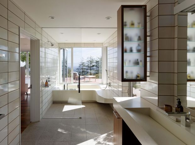 Bright contemporary bathroom with full-height white rectangular wall tiles, large grey floor tiles, a freestanding sculptural bathtub positioned by floor-to-ceiling glazing, and a long double vanity with pale solid-surface counters and timber cabinetry. A dark-framed illuminated display cabinet and large mirror add contrast. The glazing opens toward a leafy outdoor view and a terrace or balcony beyond.