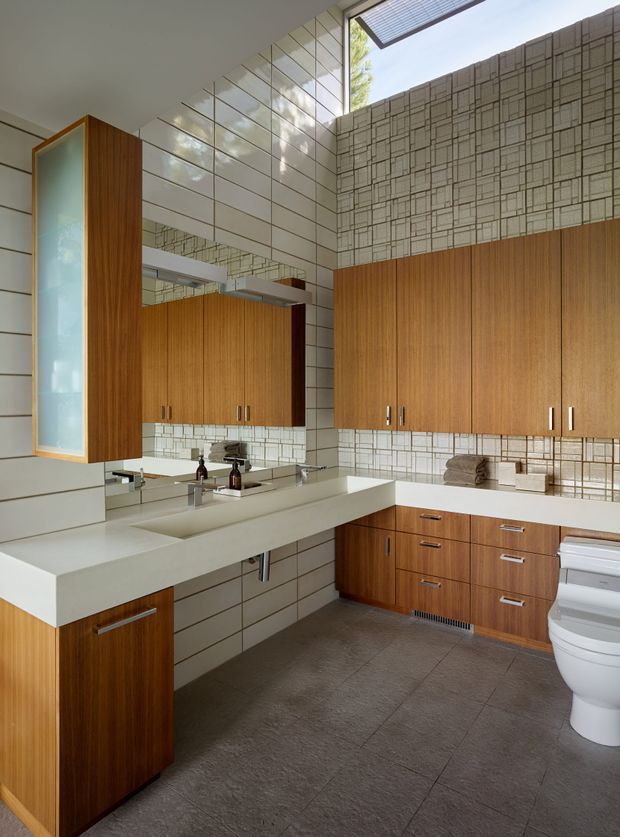 A bright, architecturally detailed bathroom with a large floating double vanity, white integrated basins, extensive mirrored surfaces, and warm vertical timber cabinetry. The walls combine large white rectangular tiles with a patterned grid-like tile feature, while the floor is finished in large dark grey tiles. A toilet is visible at right, along with a skylight and clerestory glazing that bring in natural light.