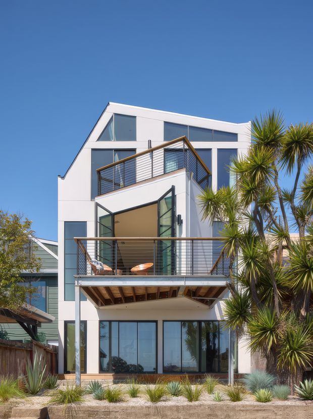 Exterior view of a contemporary multi-storey residence with a crisp white facade, extensive dark-framed glazing, angular roofline, and stacked projecting balconies with slim metal railings. The lower level opens toward a landscaped frontage, while drought-tolerant plantings and palm-like trees frame the building.