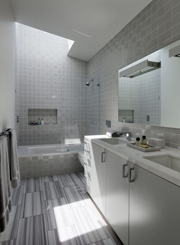 A bright contemporary bathroom with a bathtub and glass shower enclosure at the rear, a large white vanity with integrated basin and mirrored cabinet, and grey patterned wall and floor tiles. A skylight or high-level opening brings strong natural light into the space.