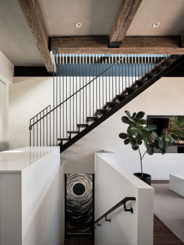 A contemporary interior stair hall viewed from an upper level, featuring a black steel open-riser staircase with slender vertical balusters, white plaster walls, exposed rustic timber ceiling beams, and a lower stair descending beside a large abstract artwork. A potted fiddle-leaf fig and a partial seating area are visible to the right. The space has a warm, refined material palette combining timber, black metal, white walls, and neutral flooring.