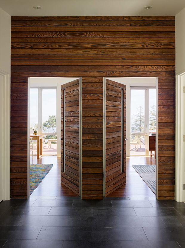 Interior view of a refined entry hall featuring a dramatic full-height dark timber feature wall with matching pivot doors and narrow metallic frames. The doors open toward bright adjoining living spaces with large windows, while the foreground has large-format dark floor tiles and recessed ceiling lights.