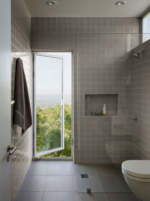 Contemporary tiled bathroom with a walk-in shower, wall niche, wall-mounted shower fittings, toilet, towel, recessed ceiling lights, and a tall opening window overlooking a leafy landscape. The room uses consistent small-format grey wall tiles and large grey floor tiles for a restrained, modern finish.