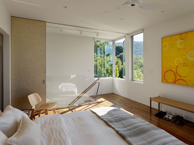 Bright contemporary bedroom with a white upholstered bed in the foreground, warm timber flooring, a woven-texture sliding door or wall panel, and expansive corner windows overlooking leafy hills. A glass balustrade and stair opening appear beside the bed, with a slim bench, chair, and colourful abstract artwork completing the room.