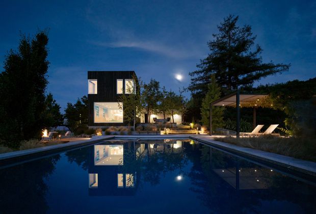 Twilight exterior view of a contemporary residential property centered on a long reflecting swimming pool. A dark, box-like two-storey pavilion with illuminated windows sits among mature landscaping, with a covered outdoor lounge and sun loungers to the right. The pool reflects the building, trees, moon, and warm architectural lighting, creating a dramatic and serene atmosphere.