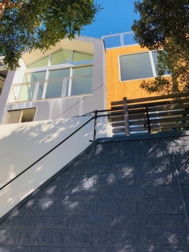 Exterior view looking upward at a contemporary multi-level residence on a sloped site. The facade combines smooth light render with a warm timber-clad upper section, large glazed windows, a rooftop or upper-level mesh balustrade, and dark horizontal metal-and-timber railings. A substantial dark masonry retaining wall and stair access dominate the foreground, with mature trees partially framing the building.