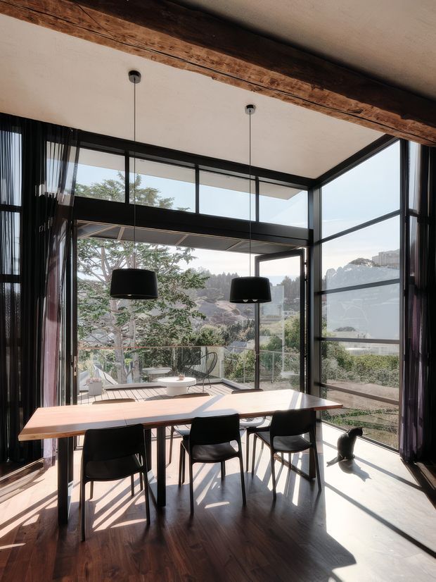 A bright, contemporary dining area with a long timber dining table and black chairs positioned beside expansive floor-to-ceiling corner glazing. Black pendant lights hang over the table, while dark curtains frame views of trees and neighbouring buildings. The room features dark timber flooring, a pale ceiling, strong natural light, and a prominent exposed timber beam across the ceiling.