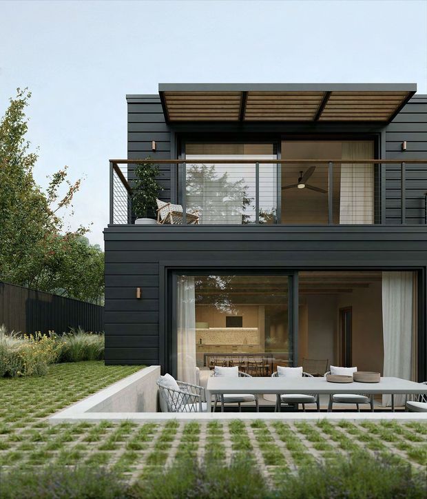 A contemporary multi-level house viewed from the rear garden, with dark horizontal cladding, extensive glazing, a projecting upper-level pergola, and a balcony with a glass-and-metal balustrade. The lower level opens onto an outdoor dining terrace, while landscaped grass and planting surround the building.