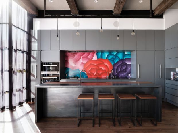 A spacious contemporary kitchen with dark grey full-height cabinetry, integrated appliances, a large island with seating for four, and a dramatic colourful abstract feature splashback. The room has exposed timber ceiling beams, black steel-framed floor-to-ceiling glazing, pendant lights, and dark timber flooring.
