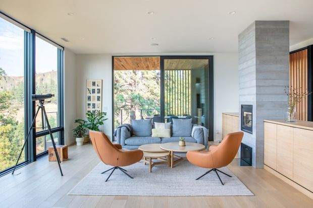 Bright, contemporary living room with floor-to-ceiling glazing overlooking a wooded landscape. The room features a grey sofa, two orange leather lounge chairs, a round timber coffee table, pale timber flooring, a large neutral rug, built-in timber cabinetry, and a concrete-look fireplace column with vertical timber screening.