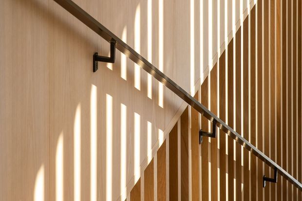 Close-up architectural view of a timber-lined stairway with a dark metal handrail and brackets. Vertical timber battens create strong rhythmic shadows and filtered sunlight across the pale wall and stair enclosure, producing a warm, graphic composition.