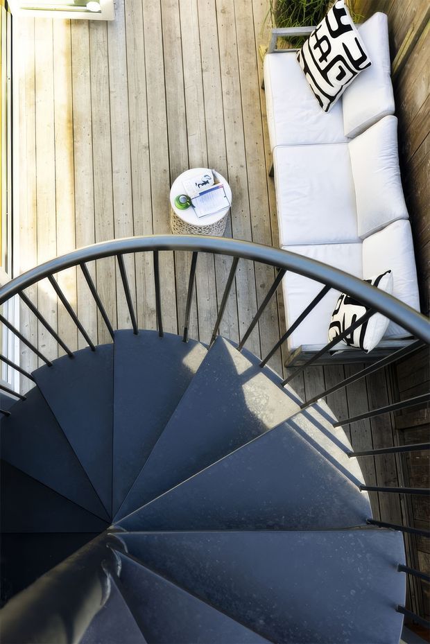 Top-down view of a compact outdoor deck or terrace with a dark spiral staircase and metal balustrade dominating the foreground. Weathered timber decking surrounds a white outdoor sofa with black-and-white patterned cushions, a small round side table, and potted greenery. The space is bright and casual with strong sunlight and shadows.