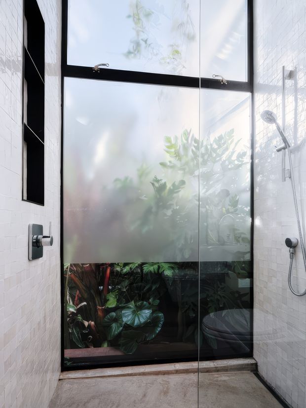 A bright contemporary bathroom with white rectangular wall tiles, a concrete-look floor, black-framed frosted glazing overlooking lush greenery, and a frameless glass shower enclosure. A black shower system and matching dark wall recess provide contrast against the pale tiled walls.