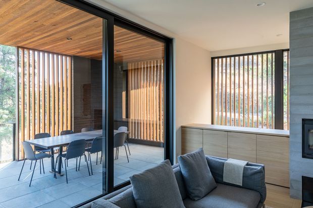 Contemporary living room opening onto a glazed dining terrace. The room features a grey sofa, light timber built-in cabinetry, a dark stone or concrete fireplace surround, pale flooring, and expansive black-framed sliding glass doors. Outside, a dining table and chairs sit beneath a warm timber-lined ceiling, with vertical timber screening providing privacy and filtered light.