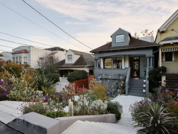 Street-facing view of a compact, traditional timber-clad house with a steep gabled roof, front dormer, covered porch and central stair entry. The dark grey-blue facade is surrounded by a densely planted, drought-tolerant garden with concrete paths and retaining walls. Neighboring homes and overhead utility wires are visible in the suburban streetscape.