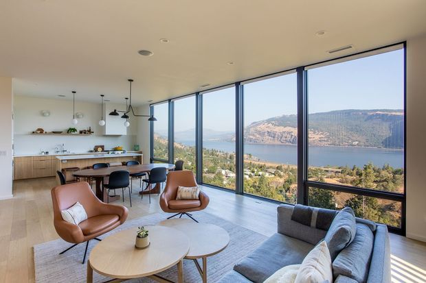 A bright, open-plan living and dining space with a contemporary kitchen at the rear and expansive floor-to-ceiling glazing overlooking a lake and rugged mountain landscape. The room features timber flooring, a pale neutral ceiling and walls, a minimalist kitchen with a long island, pendant lights, a round dining table, sculptural leather lounge chairs, a grey sectional sofa, and nested oval coffee tables. The glazing creates a strong indoor-outdoor connection and fills the space with natural light.