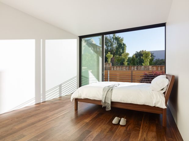 A bright, minimalist bedroom with a low-profile solid timber bed, white bedding, and a warm dark timber floor. Large black-framed sliding glass doors open to a timber privacy screen and leafy outdoor setting. Strong afternoon sunlight creates crisp geometric shadows across the white walls and floor.