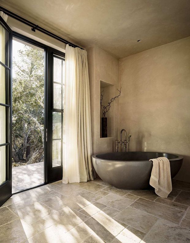 A serene, warm-toned bathroom featuring a large freestanding oval bathtub with a dark interior, brushed metal floor-mounted faucet, and a recessed wall niche containing decorative branches. Full-height black-framed glazed doors open to an outdoor terrace and trees, softened by cream curtains. The room has a textured plaster finish and large irregular stone-look floor tiles, with strong natural light creating long shadows.