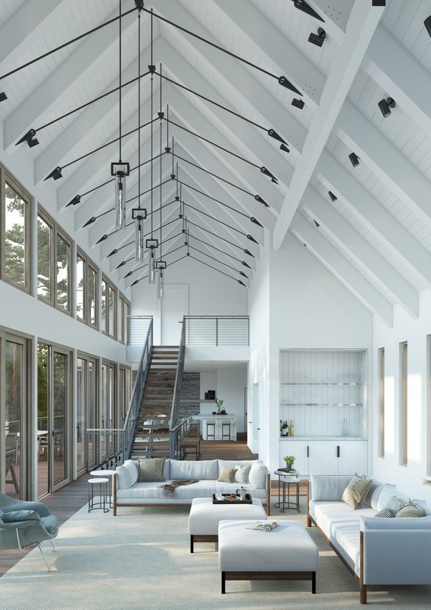 A bright, expansive double-height living room with a dramatic white pitched timber ceiling and exposed structural beams. Repeated black pendant lights hang along the long vaulted space, while extensive floor-to-ceiling glazing lines one side. The room contains multiple pale upholstered sofas and armchairs, low tables, a large rug, and a contemporary open stair leading to a mezzanine. A built-in white shelving and bar area is visible at the rear.