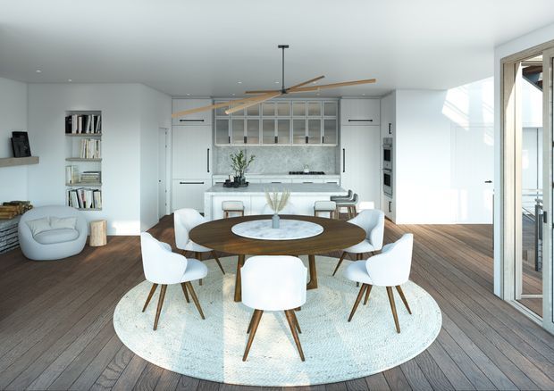 Bright open-plan interior featuring a dining area with a round dark timber table and white upholstered chairs on a large round rug, opening toward a minimalist white kitchen with integrated cabinetry, marble-look splashback, island bench, and bar stools. Warm timber flooring, white walls, expansive glazing, and a large timber-bladed ceiling fan create an airy contemporary setting.