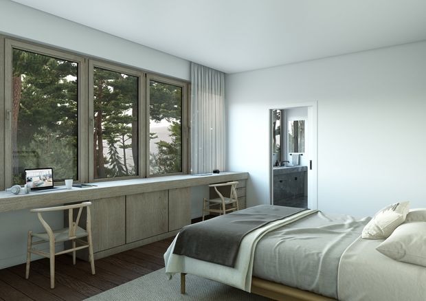 A bright, calm contemporary bedroom with a low built-in timber window seat and desk running beneath large framed windows, two light timber chairs, a neatly made bed with layered neutral bedding, and a compact bathroom visible through the open doorway. The room uses white walls, dark timber flooring, muted grey-green cabinetry, and full-height curtains, with views of mature trees outside.