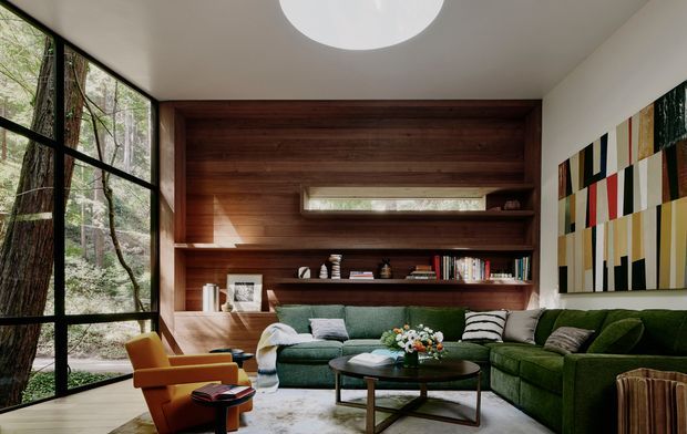 A sophisticated living room with floor-to-ceiling black-framed glazing overlooking a wooded setting, a dark timber built-in shelving and feature wall, a deep green sectional sofa, mid-century-style lounge chairs, a low coffee table, and colourful abstract wall art. A circular ceiling skylight brings additional daylight into the warm, refined interior.