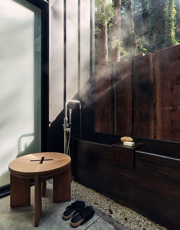 A moody, compact bathing space with a dark freestanding bathtub positioned beside a large glazed opening and timber privacy screen. A handheld shower rises beside the tub, with steam visible in the air. The room features dark timber wall and tub surfaces, a grey concrete-like floor, a small round timber stool or table, and a narrow strip of decorative gravel. Dense greenery is visible beyond the glazing, creating an indoor-outdoor spa atmosphere.