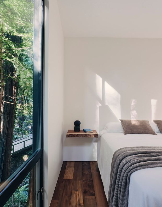 A bright, minimalist bedroom with a white bed, textured taupe pillows, and a grey throw. A compact floating bedside table with a warm timber top sits beside the bed, while dark timber flooring and a full-height black-framed window provide contrast. The window overlooks dense greenery and a raised outdoor walkway or deck. Sunlight creates geometric shadows across the white wall.
