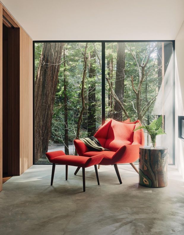 A refined sitting area with a sculptural red lounge chair and matching ottoman positioned beside expansive black-framed glazing overlooking dense woodland. The room features a polished concrete floor, warm vertical timber wall panelling and joinery, white walls, and a metallic cylindrical side table with a fern. The composition is calm and nature-connected, with strong indoor-outdoor visual continuity.