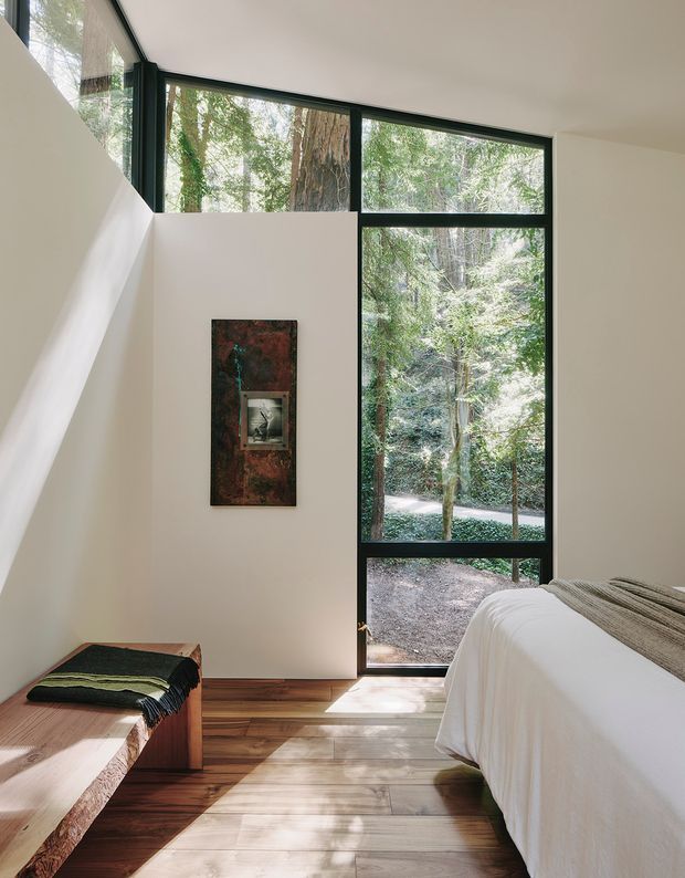 A serene, minimalist bedroom with full-height black-framed glazing overlooking dense woodland. The room has warm timber flooring, white walls and ceiling, a simple white-covered bed, and a low timber bench with a dark striped throw. A narrow sculptural wall artwork adds a muted accent, while strong natural light and the forest view create a calm connection to nature.