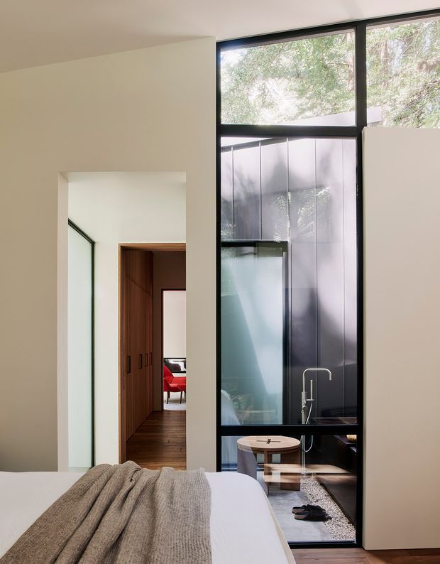 A modern bedroom viewed toward a tall black-framed glazed partition and adjoining interior spaces. The foreground shows a neatly made bed with a textured taupe throw. Through the glazing is a compact sitting area with a round timber table and chairs, while an open passage reveals dark timber built-in cabinetry and a glimpse of a red chair. The room is bright and understated, with pale plaster walls, dark-framed glazing, timber flooring and strong visual connection to leafy trees outside.