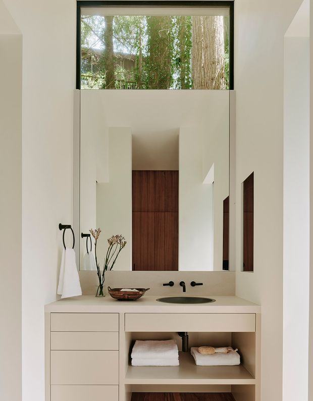 A refined minimalist bathroom vanity beneath a high clerestory window. The white built-in vanity has flat-front drawers, an open lower shelf, a pale solid-surface benchtop, an integrated dark oval basin, and black wall-mounted tapware. A large frameless mirror reflects the room, while warm vertical timber wall panelling appears in the rear alcove. Natural greenery is visible outside the window.