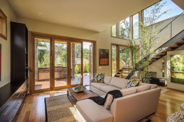 Warm, light-filled contemporary living room with a large cream sectional sofa, timber flooring, dark built-in media cabinetry, and expansive timber-framed glazing opening to an outdoor deck. A sculptural open-riser staircase and double-height glazing create a strong vertical architectural feature, with indoor planting and artwork adding natural character.