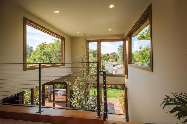 A bright contemporary landing or upper-level hallway overlooking a lower living area, with warm timber-framed corner windows, cream walls, recessed ceiling lights, and a black metal-and-glass balustrade. Lush greenery and a residential neighbourhood are visible outside, while an open stairwell and timber flooring create a spacious, connected interior.