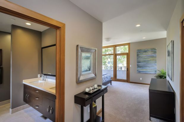 Interior view from a hallway or landing looking toward a carpeted living area, with an open bathroom visible on the left. The bathroom features a dark floating vanity, white benchtop, large mirror, and tiled floor. Timber door frames, neutral painted walls, recessed ceiling lights, a narrow console table, framed artwork, and a dark sideboard define the contemporary transitional interior.
