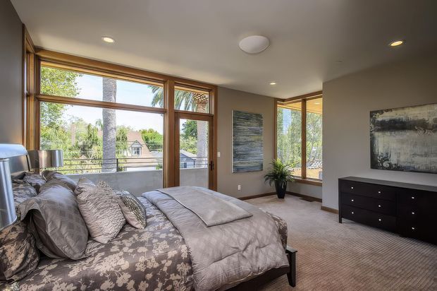 A bright, contemporary bedroom with a bed positioned beside expansive timber-framed windows and glazed doors. The room has neutral taupe walls, soft patterned bedding, carpeted flooring, a dark low dresser, recessed ceiling lights, and abstract wall art. Trees and neighboring houses are visible outside through the glazing.