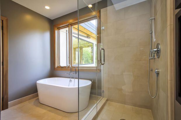 A bright, refined bathroom featuring a freestanding white bathtub beside a large timber-framed window and a fully tiled walk-in glass shower. Beige stone-look floor and wall tiles are paired with dark taupe painted walls, recessed ceiling lighting, and chrome shower fittings.
