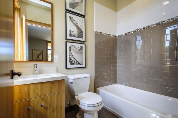 A compact, warmly styled bathroom with a timber vanity, white basin, toilet, and bathtub. The walls feature glossy grey subway tiles with a decorative circular border, while the vanity wall is painted a soft neutral tone. A large timber-framed mirror and three framed black-and-white architectural prints add visual interest. The room is bright, clean, and moderately sized.