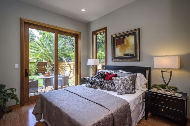 A warmly styled bedroom with a queen or king bed, grey textured bedding, patterned and accent cushions, dark bedside furniture, table lamps, framed artwork, and timber-framed glazed doors opening to a small outdoor terrace or garden. Natural greenery is visible through the glazing, while recessed ceiling lighting provides additional illumination.