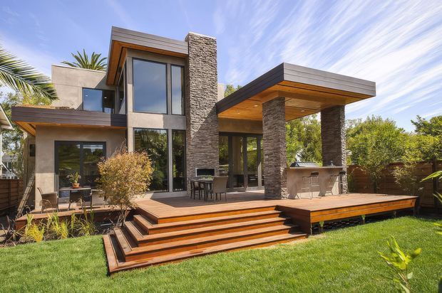 Contemporary two-storey residential home with a strong indoor-outdoor connection, featuring large dark-framed glazing, grey textured stone-clad columns and chimney, smooth concrete-toned walls, and projecting flat roof forms. A broad timber deck with stepped access surrounds an outdoor dining area and covered outdoor kitchen or barbecue zone. The landscaped backyard includes lawn and tropical planting.