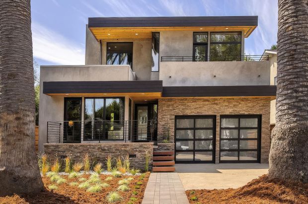 Two-storey contemporary residence photographed from the front, with flat roof planes, projecting balconies, dark metal-framed glazing, warm timber soffits, textured stone veneer, pale rendered walls, and an integrated double garage. A paved path leads through landscaped planting toward the entry, framed by mature palm trees.