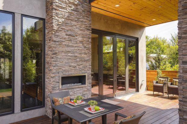 A bright, contemporary outdoor living and dining area framed by stacked-stone columns and walls. The space features a built-in linear fireplace, dark-framed floor-to-ceiling glazing and folding doors, a warm timber-lined soffit, timber decking, and a dining table with woven chairs. A secondary seating area sits beyond the open doors on the right, enclosed by timber fencing and planting.