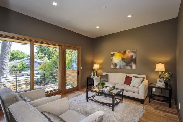 A warm, contemporary living room with dark taupe walls, recessed ceiling lights, and timber-framed floor-to-ceiling glazing opening to a balcony or deck. The room contains a light upholstered sofa and armchairs, a black metal-and-glass coffee table, side tables with lamps, a large pale rug, timber flooring, and abstract artwork.