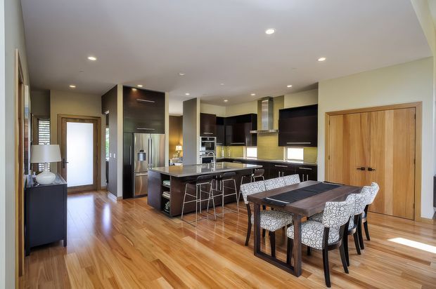 Open-plan kitchen and dining area with warm timber flooring, dark espresso cabinetry, a large central island with bar seating, stainless-steel appliances, and a timber dining table surrounded by patterned upholstered chairs. Recessed ceiling lighting and light walls create a bright, contemporary interior.