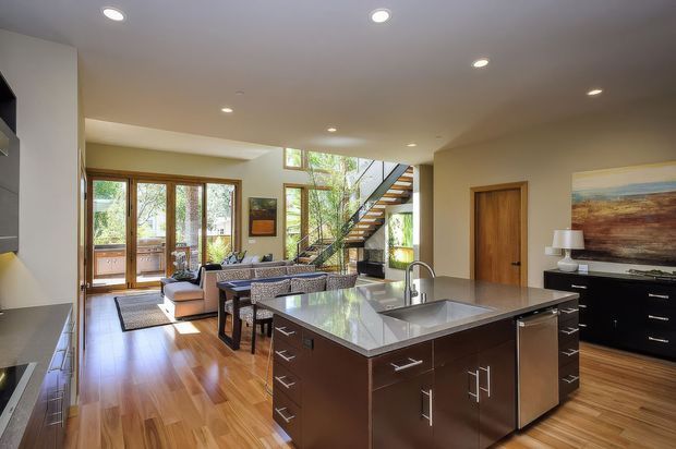 Open-plan contemporary kitchen and living area with a large dark wood island, pale stone benchtop, stainless-steel appliances, polished timber flooring, recessed ceiling lights, and warm timber-framed glazing. A sitting area and open-riser staircase are visible beyond the kitchen.