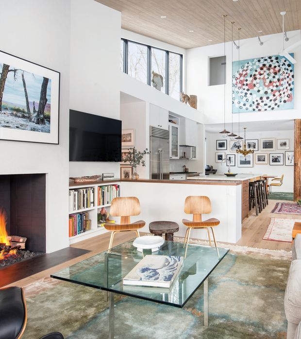 A bright, double-height open-plan living and kitchen area with a dramatic fireplace, wall-mounted television, built-in bookcase, glass coffee table, sculptural wood chairs, and a kitchen island. White walls and extensive pale timber ceiling panelling create an airy contemporary interior, while framed artwork and a gallery wall add visual interest. A mezzanine-like upper level and tall glazing emphasize the vertical scale.