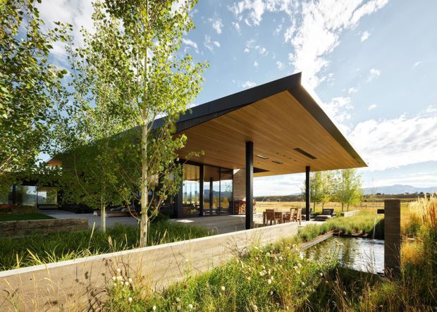 A contemporary single-storey pavilion-like home or outdoor entertaining structure set within a landscaped rural or semi-rural site. The building has a dramatic asymmetrical pitched roof with a deep timber-lined soffit, dark slender structural posts, extensive floor-to-ceiling glazing, and a covered outdoor dining area opening toward a narrow landscaped water feature. Native-style planting, stone retaining walls, and expansive views contribute to an integrated indoor-outdoor setting.