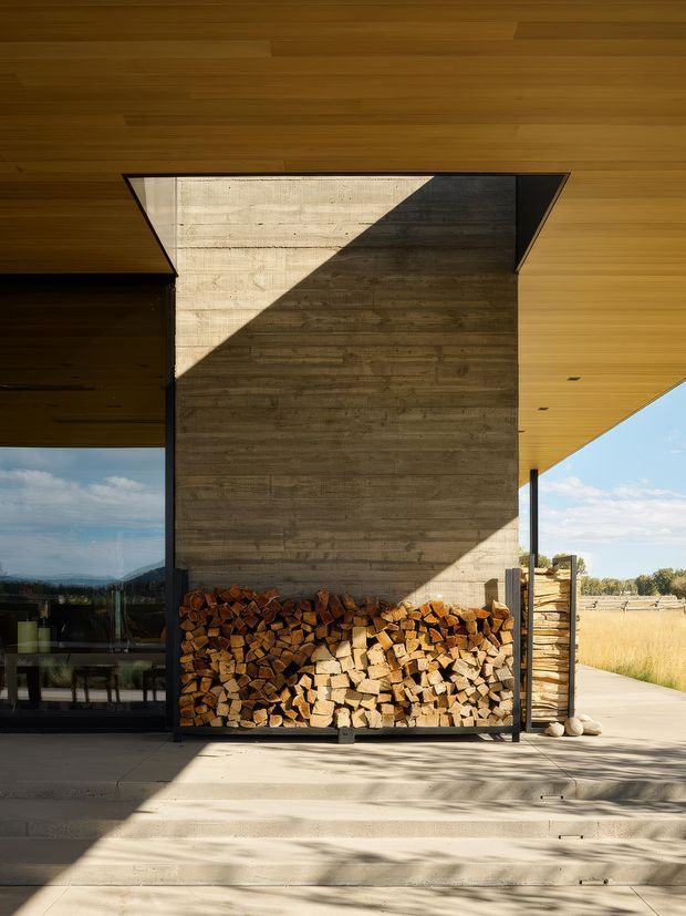 A contemporary rural home exterior or covered outdoor living area featuring a large exposed concrete feature wall, a built-in firewood storage bay filled with split logs, dark metal framing, and extensive warm timber soffit cladding. Glazed openings reveal an interior dining area, while the open side looks across a dry rural landscape. Strong geometric sunlight and shadow create a dramatic architectural composition.