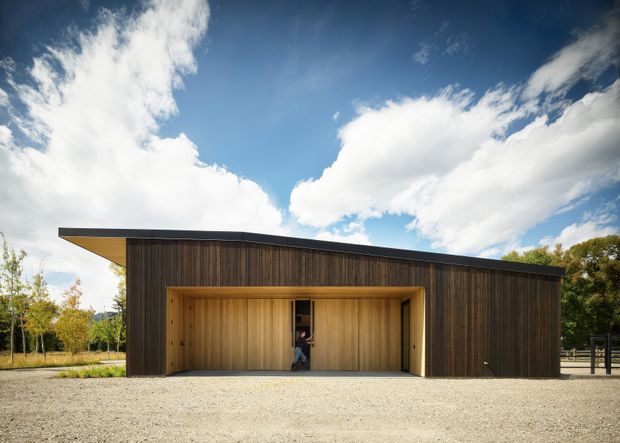 A low, single-storey contemporary building with a long dark timber-clad facade and a dramatic projecting flat roof. A recessed central entry is framed in warm timber, opening onto a broad gravel forecourt. The minimalist composition is set against a bright blue sky with trees visible around the edges.