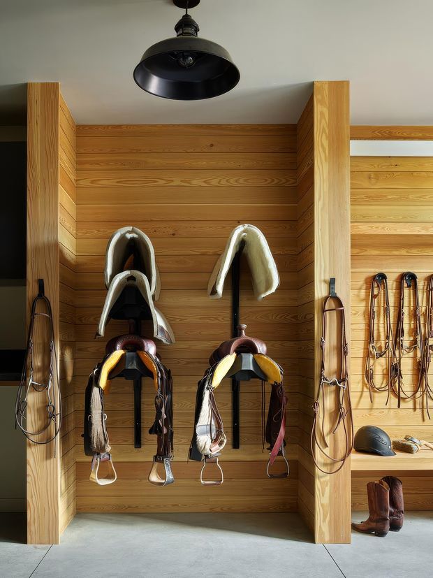 A neatly organised equestrian tack storage area with several saddles, bridles, riding helmets and boots displayed against warm horizontal timber wall panelling. Vertical timber dividers frame the recessed storage bays, while a dark industrial-style pendant light hangs above. The concrete floor and exposed black hooks contrast with the warm wood joinery.